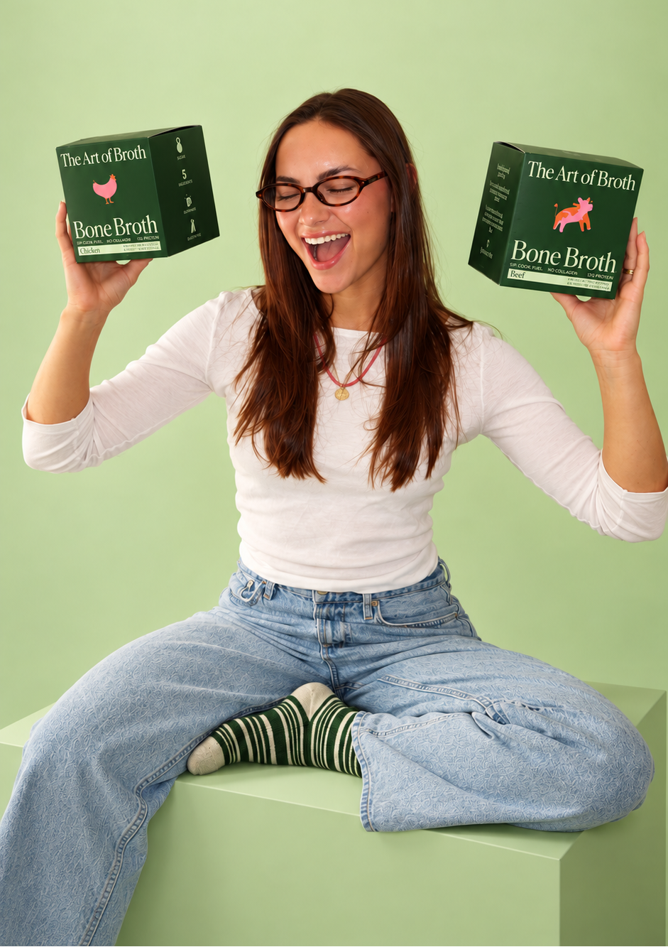 A woman with glasses sits cross-legged on a light green pedestal, holding two boxes of 'The Art of Beef Bone Broth' on each side. She is wearing a white long-sleeve top, light blue jeans, and green-and-white striped socks against a solid light green background.